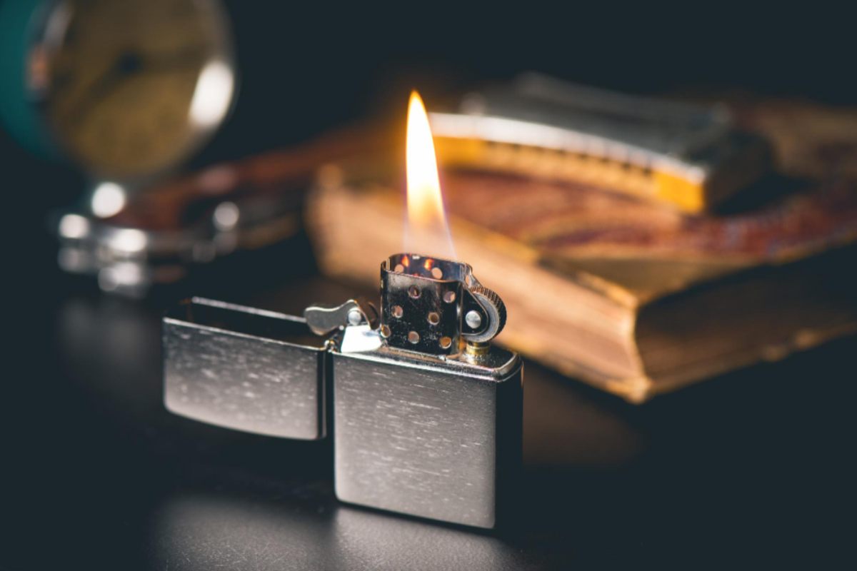 Briquet tête de mort chromé ouvert et allumé, posé sur bureau en bois avec montre à gousset et livre ancien